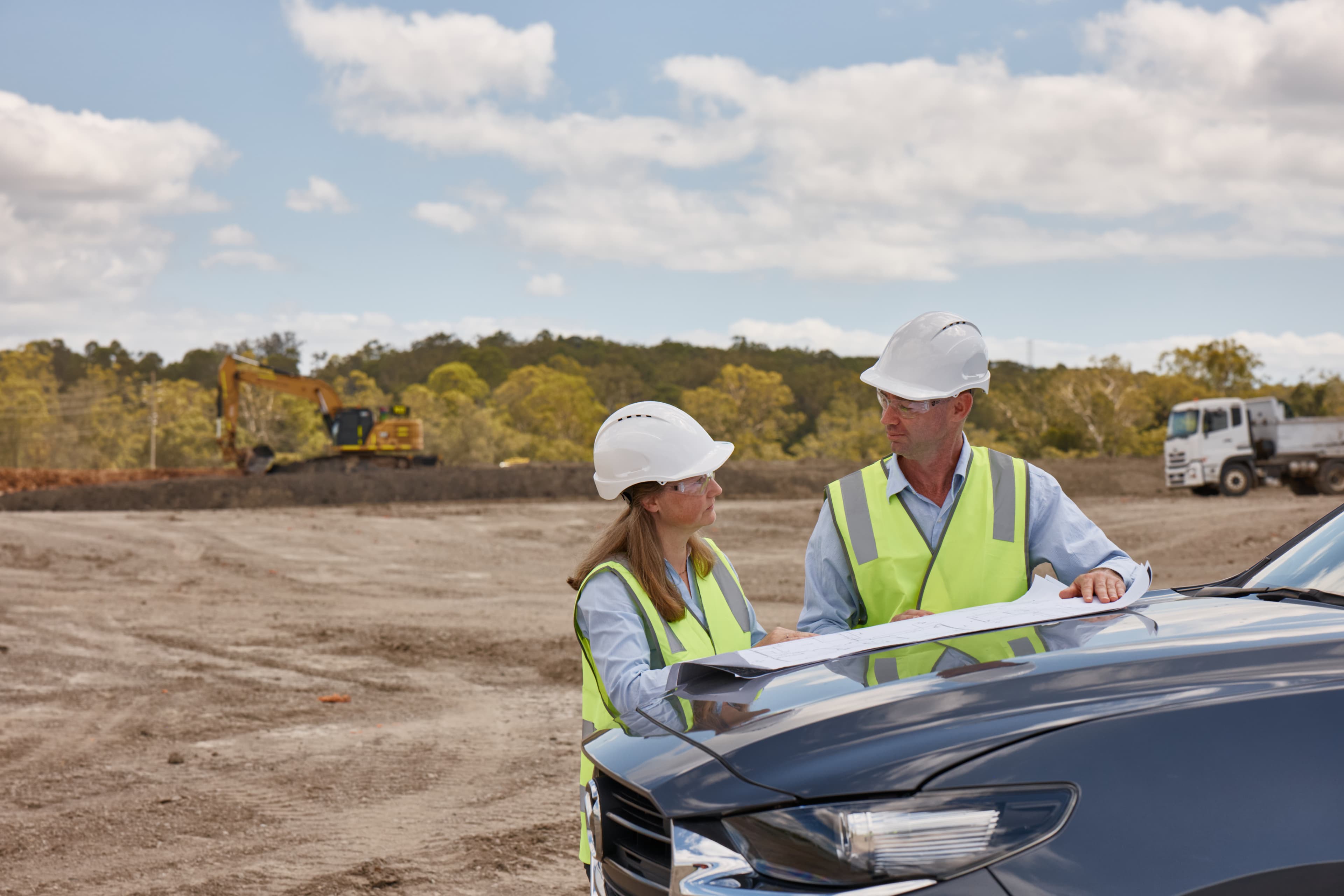 Two construction workers looking at plans on the hood of a car at a construction site