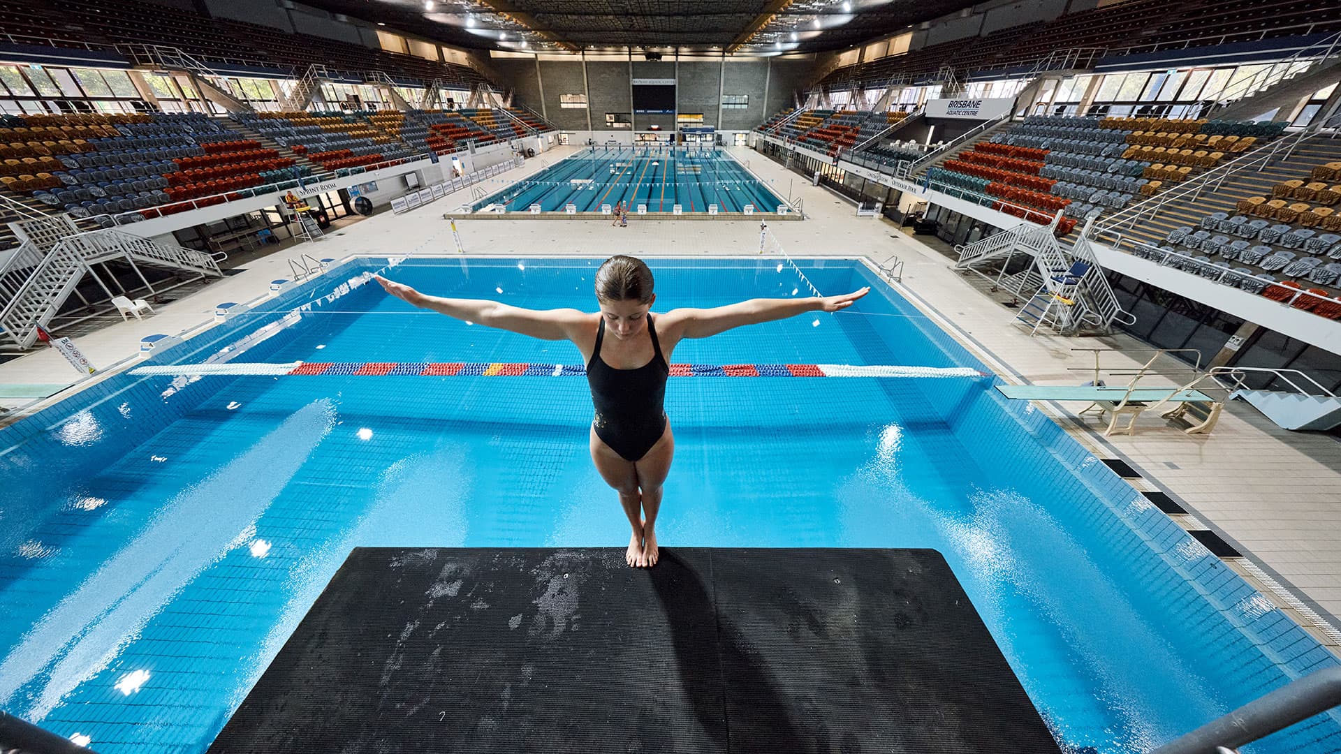 A diver stands on a platform with arms outstretched above an indoor pool