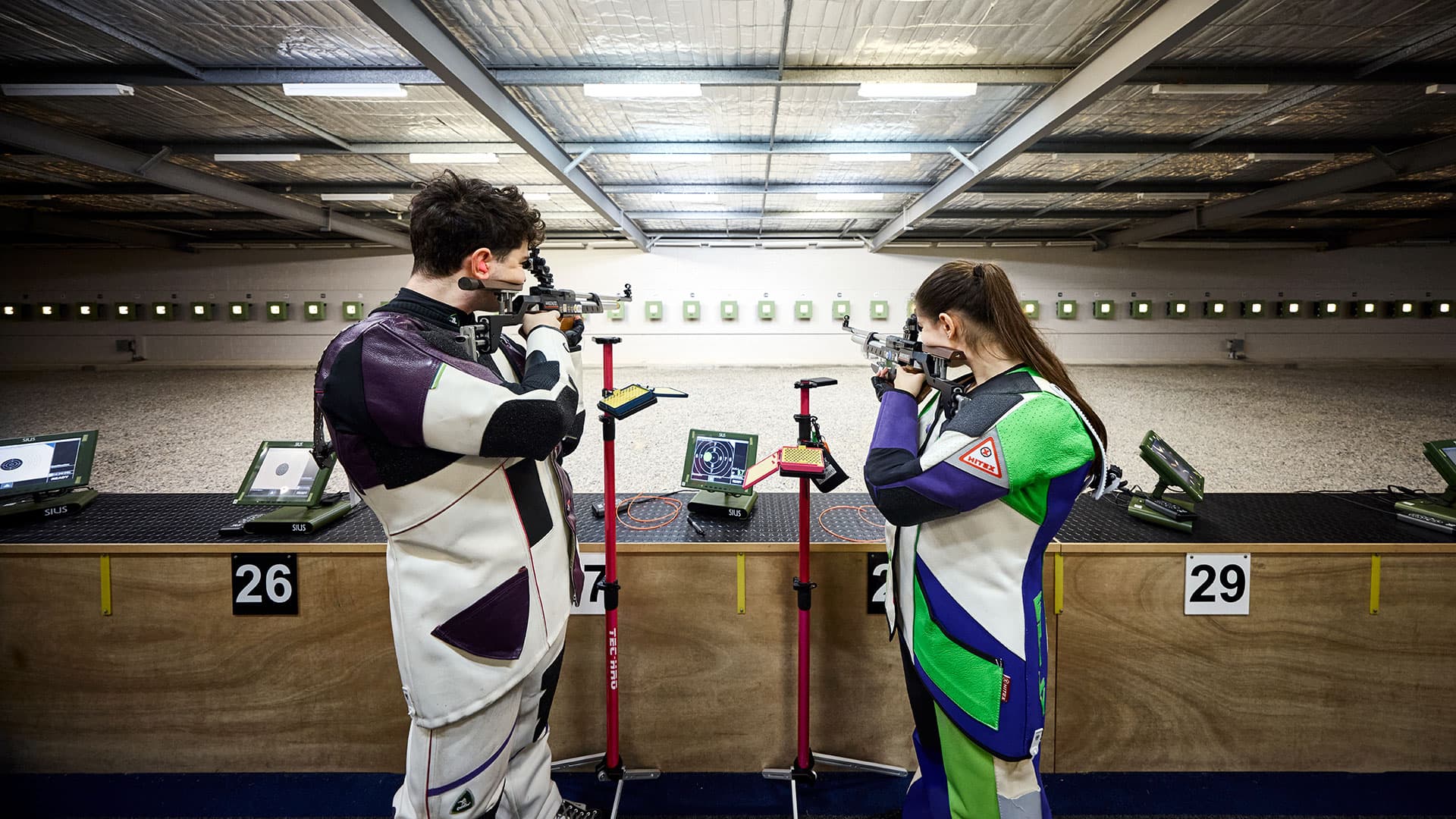 Two people at an indoor shooting range aiming rifles at targets