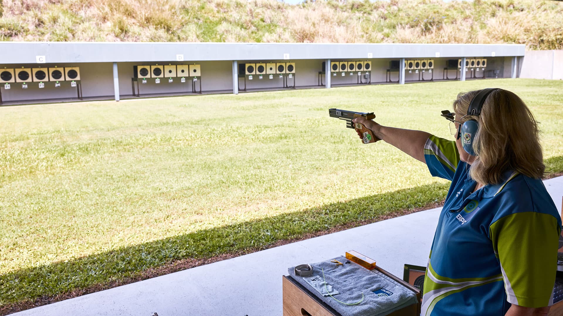 A person stands at an outdoor shooting range aiming a pistol toward distant targets