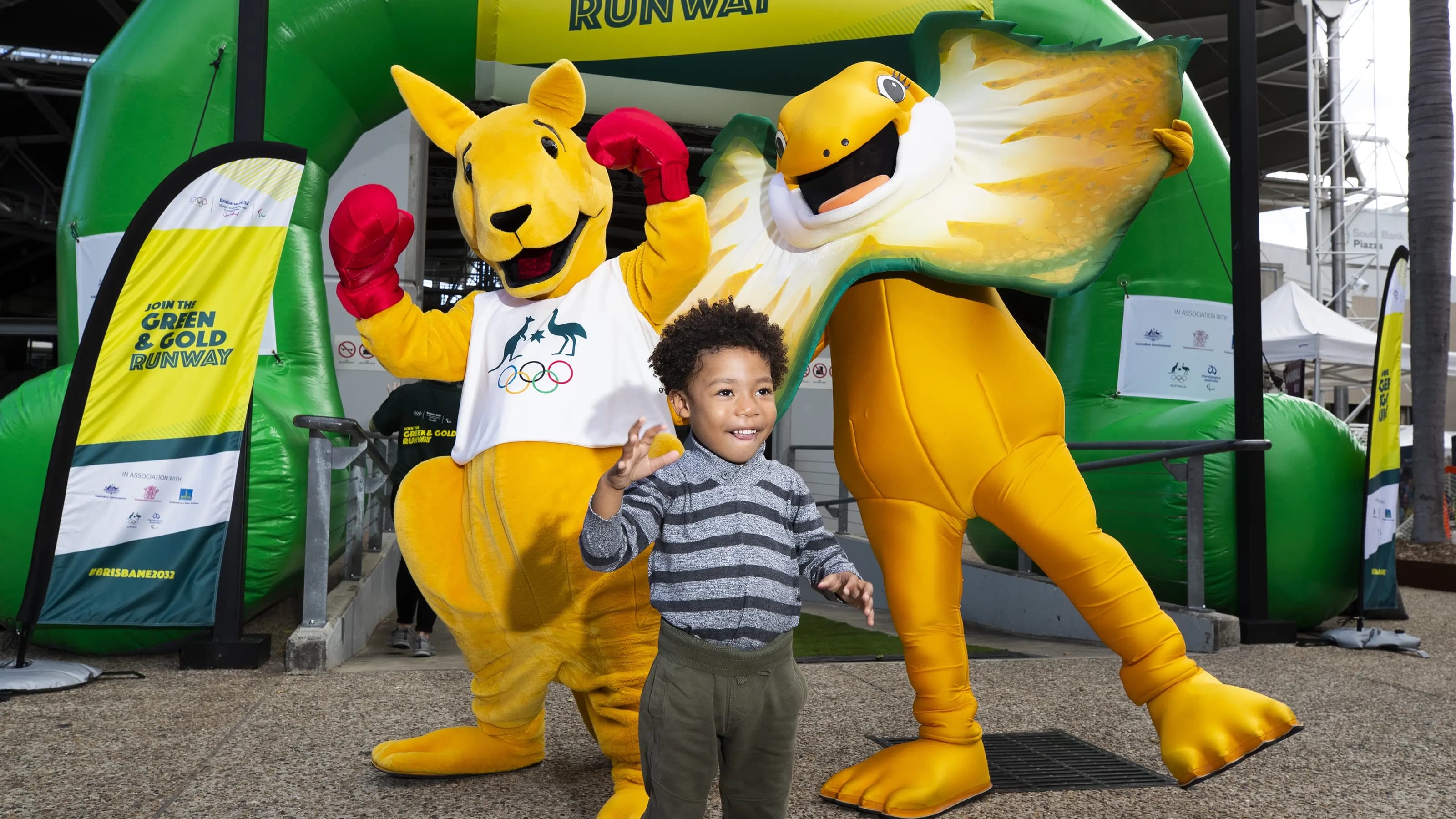 Boxing Kangaroo and Lizzie, a frill-necked lizard stand in front of a 'join the green and gold runway' blowup banner, with a young boy in front of them